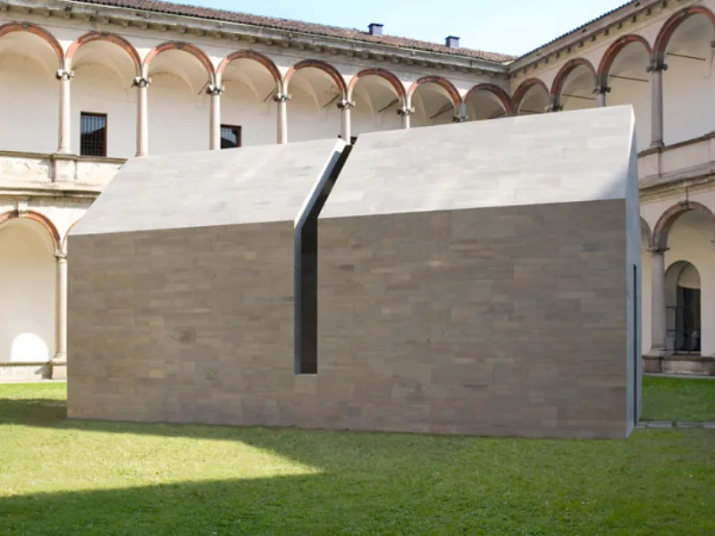 casa in pietra grigia con apertura verticale nel centro all'interno di chiostro con prato